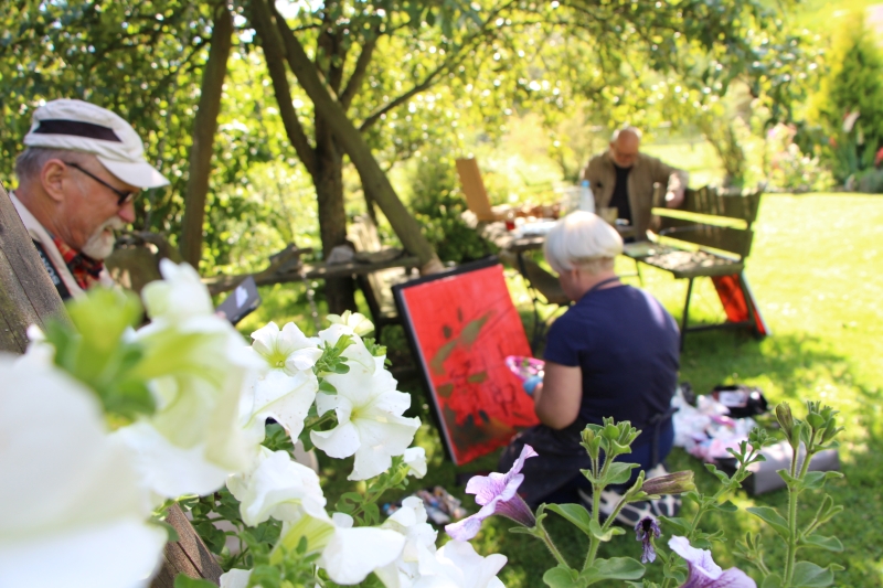Plener, kobieta maluje obraz na sztaludze, mężczyzna siedzi na ławeczce, dookoła drzewa, krzewy, zieleń.