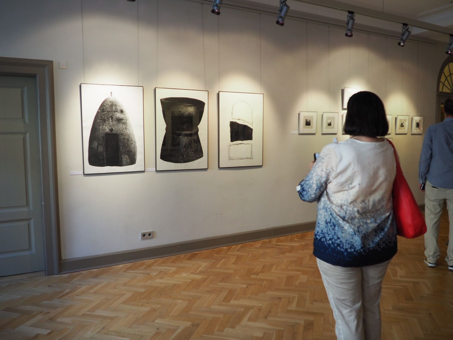 Na zdjęciu widać ścianę sali wystawowej na której wiszą trzy duże obrazy i kilka małych prezentujących sztukę współczesną. Na pierwszym planie stoi tyłem do obiektywu kobieta przyglądająca się pracom.
