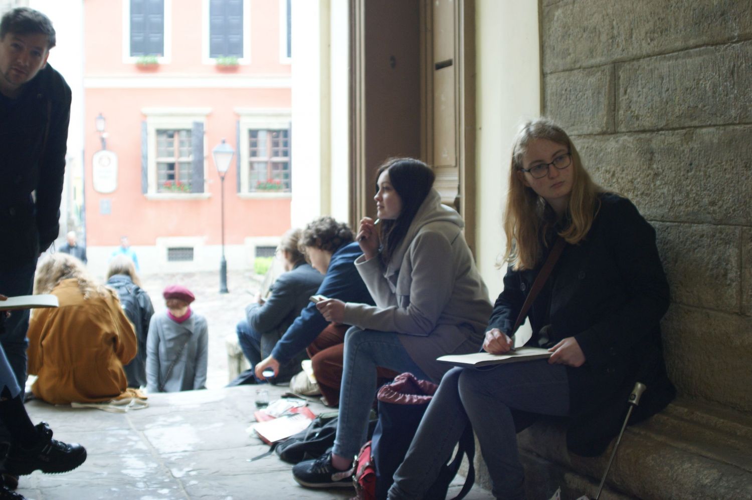 Grupa uczniów siedząca na wąskiej uliczce i rysująca na kolanach budynki znajdujące się przed ulicą.