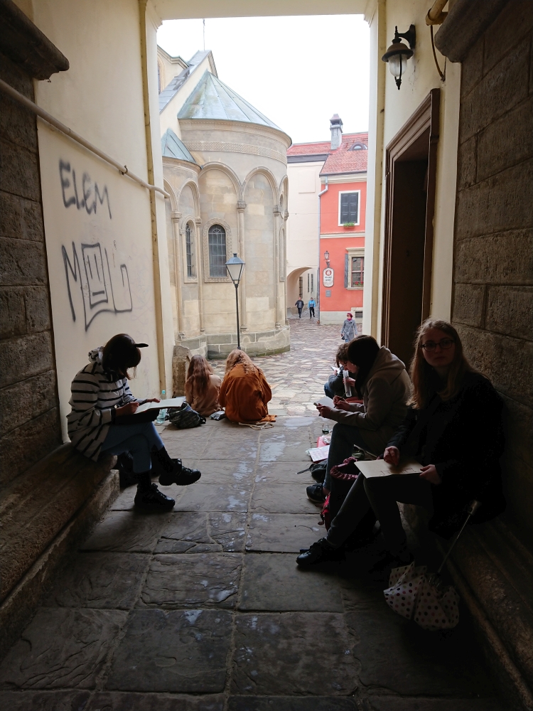 Grupa uczniów rysujących na siedząco budynki które widzą przed sobą, siedzą w wąskiej uliczce.