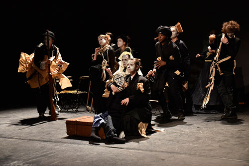 Zdjęcie przedstawia scenę teatru na której widać grupę aktorów w różnych pozach, część stoi, cześć siedzi, inni nachylają się nad innymi osobami